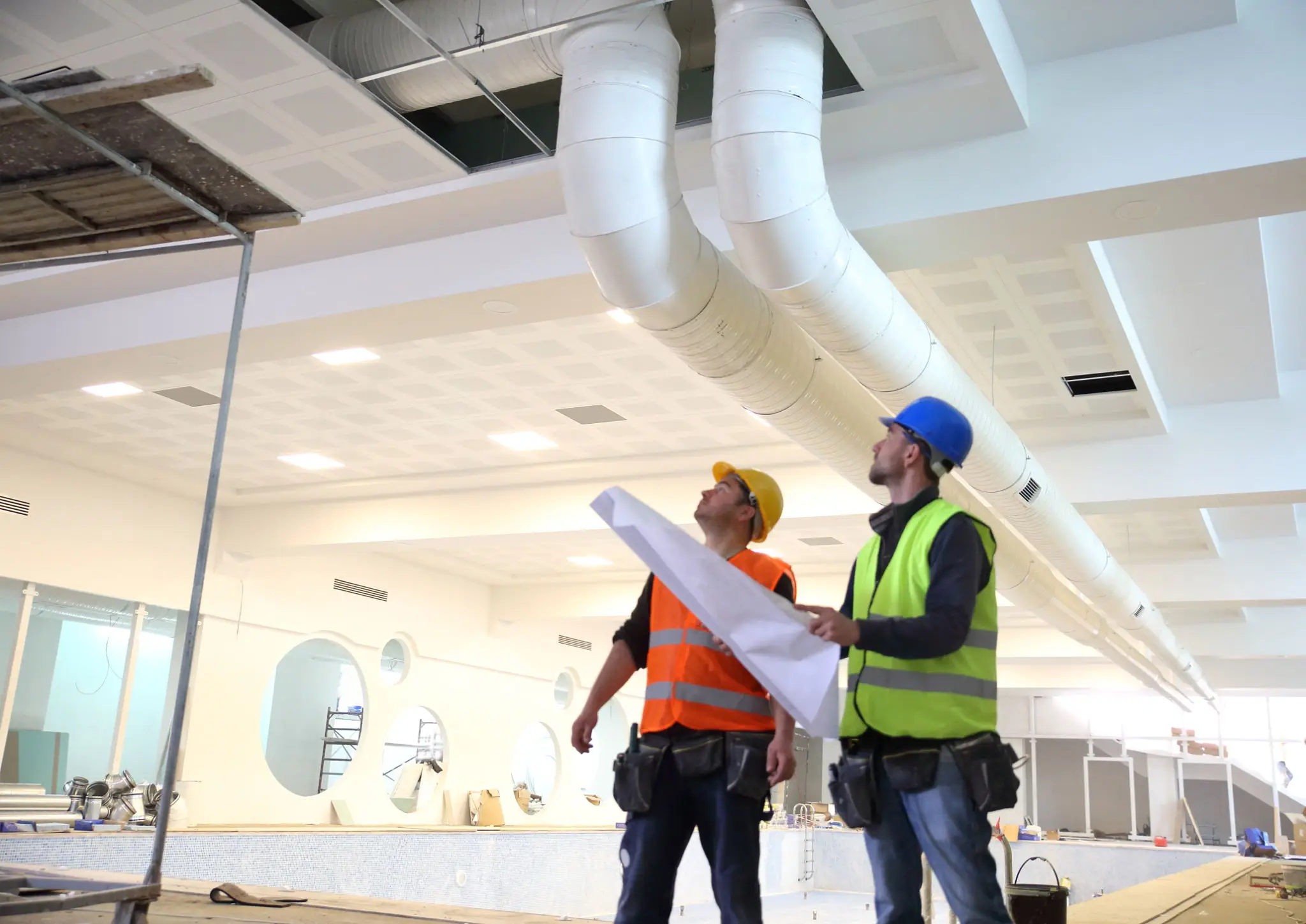 Two construction workers inspecting ceiling pipes inside a building.